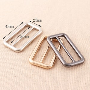 May include: Three metal belt buckle sliders in silver, gold, and black. The sliders are rectangular with rounded corners and measure 4.3 cm by 3.5 cm.