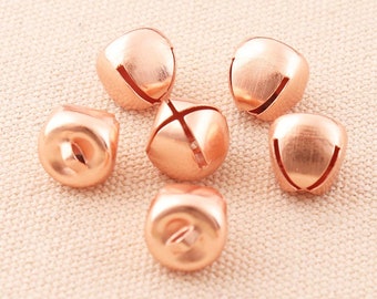 Puede incluir: Un grupo de seis pequeñas campanillas de color oro rosa. Las campanillas están esparcidas sobre una superficie de tela beige claro.
