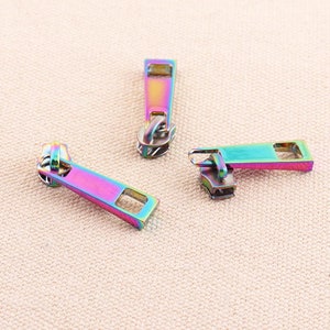 May include: Three iridescent zipper pulls in a rainbow color scheme. The pulls are made of metal and have a rectangular shape with a small hole at the top for attaching to a zipper.