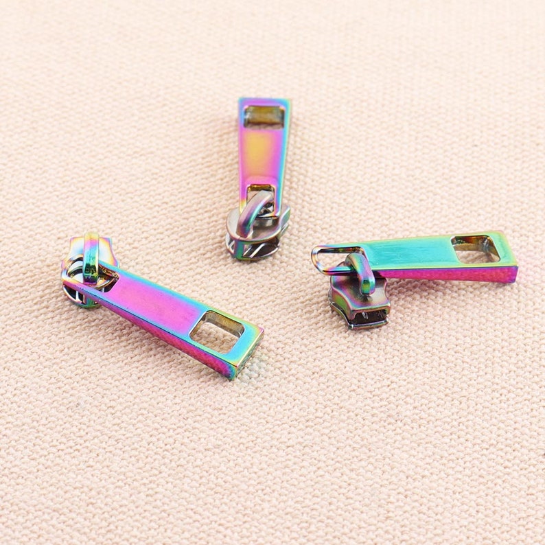 May include: Three iridescent zipper pulls in a rainbow color scheme. The pulls are made of metal and have a rectangular shape with a small hole at the top for attaching to a zipper.