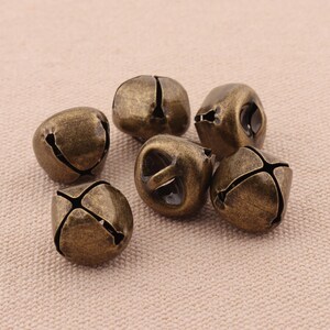 May include: Six antique bronze-colored bells with a weathered finish. Each bell has a cross-shaped slit design and a small opening at the top. The bells are clustered together on a neutral-toned surface.