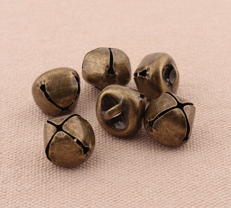 May include: Six antique bronze-colored bells with a weathered finish. Each bell has a cross-shaped slit design and a small opening at the top. The bells are clustered together on a neutral-toned surface.