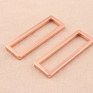 May include: Two rose gold metal rectangular rings, each with a square opening in the center. The rings are lying flat on a light brown textured surface.