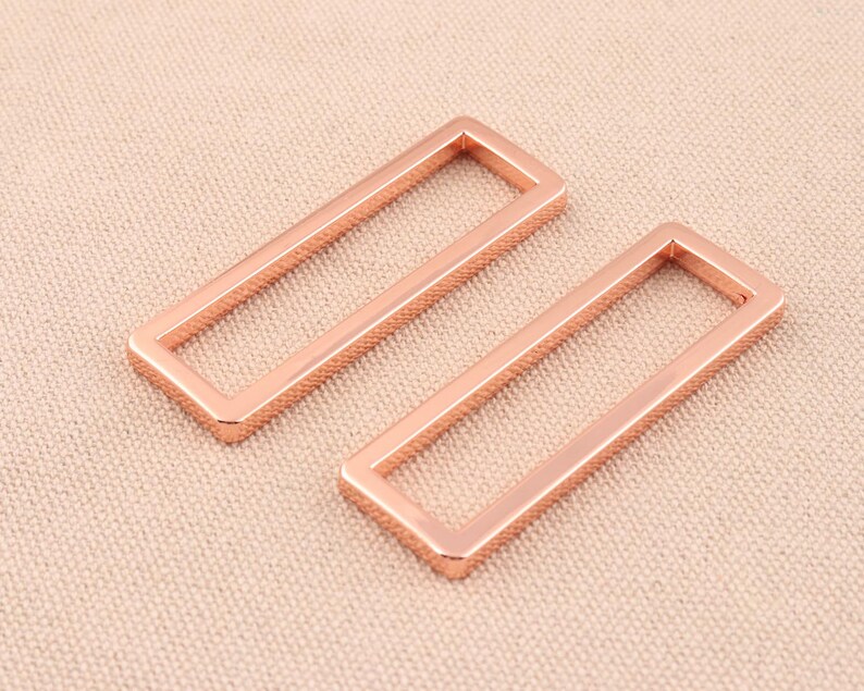 May include: Two rose gold metal rectangular rings, each with a square opening in the center. The rings are lying flat on a light brown textured surface.