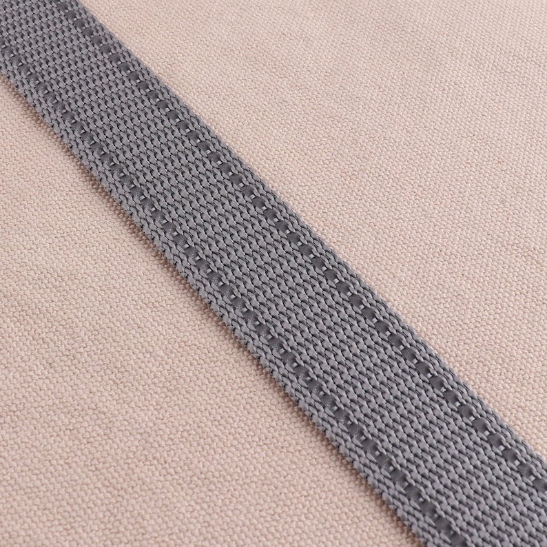 May include: A close-up of a gray woven strap with a stitched edge. The strap is on a light brown fabric background.