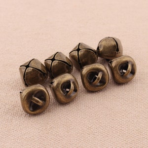 May include: A collection of eight antique bronze-colored bells. Each bell has a square shape with a cross-shaped slit on top and a loop for hanging. The bells are arranged on a neutral beige surface.