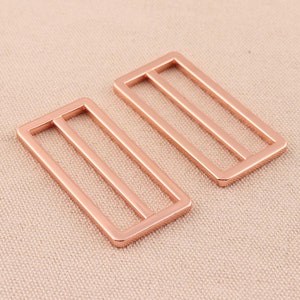 Pode incluir: Duas fivelas deslizantes de metal na cor ouro rosa. Cada fivela tem uma forma retangular com três barras paralelas.