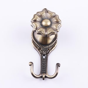May include: Antique gold-toned metal hook with a decorative flower design. The hook has two prongs and is designed for hanging items.