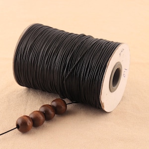 May include: A spool of black waxed cord with a label that reads "1/2 mm". The cord is wrapped around a cardboard spool with a white label. A string of wooden beads is threaded through the cord.