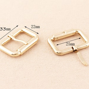 May include: Two gold metal belt buckles. The buckle on the left measures 8.4 cm by 5.6 cm. The buckle on the right measures 6.4 cm by 2.5 cm.