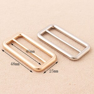 May include: Two metal rectangular slider buckles, one gold and one silver. The gold buckle measures 48mm by 40mm by 25mm. The silver buckle is slightly larger.