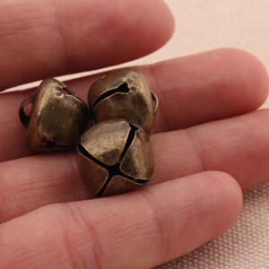 May include: Three small, antique brass-colored bells. Each bell has a rounded, square shape with a slit design and a small loop at the top. The bells are held in a person's hand.