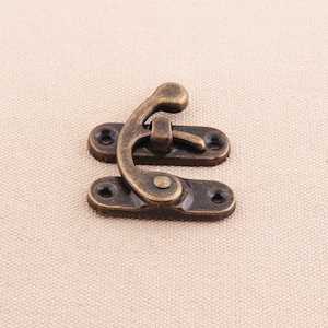 May include: An antique-style bronze-colored latch, featuring a curved hook and two rectangular base plates with screw holes. The latch is designed for securing small boxes or decorative items.