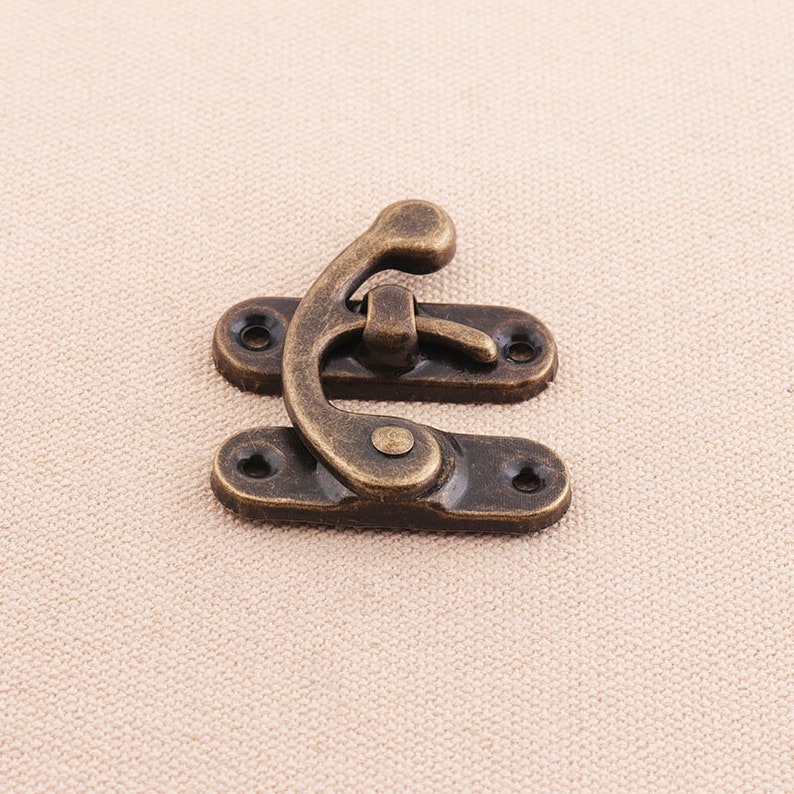 May include: An antique-style bronze-colored latch, featuring a curved hook and two rectangular base plates with screw holes. The latch is designed for securing small boxes or decorative items.