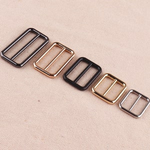 May include: Five metal slider buckles in different sizes. The buckles are arranged in a row from largest to smallest. The buckles are black, gold, and silver.