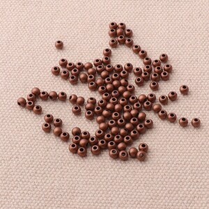 Peut inclure: Un ensemble de petites perles rondes de couleur cuivre, dispersées sur une surface de ton neutre. Chaque perle est percée d'un petit trou en son centre, adapté à l'enfilage. Ces perles sont idéales pour la création de bijoux et les projets d'artisanat.