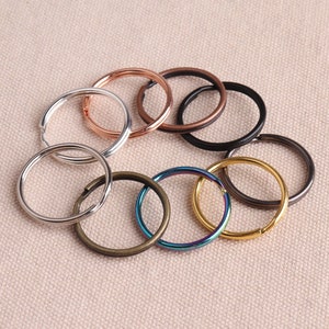 Könnte beinhalten: Sortiment an Schlüsselringen in verschiedenen Farben und Ausführungen, darunter Silber, Gold, Schwarz und Bronze. Die Schlüsselringe sind in einem Kreis auf einem beigefarbenen Stoffhintergrund angeordnet.