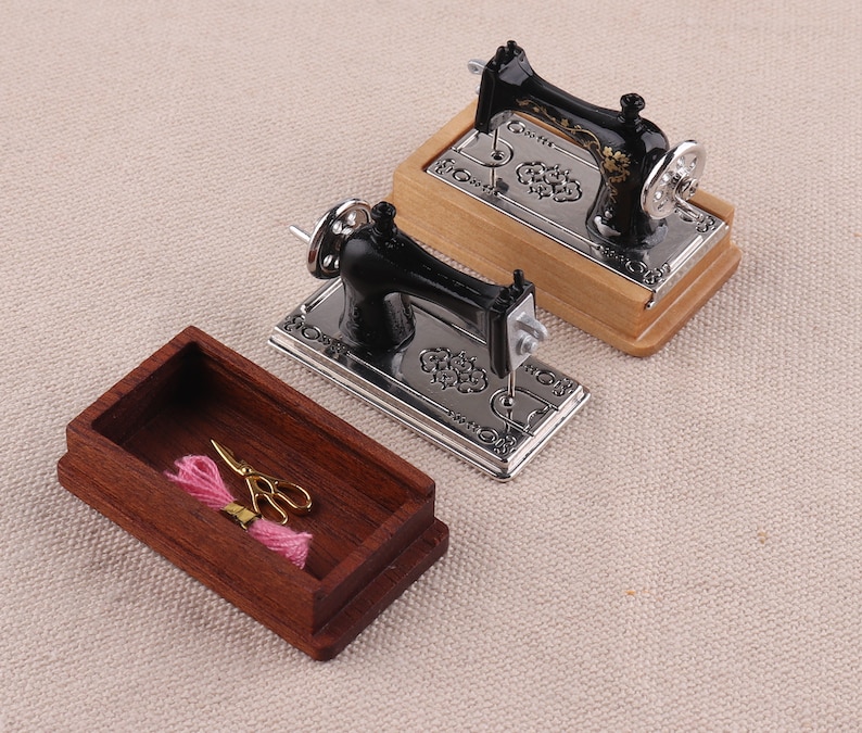 May include: Two miniature black and silver sewing machines with ornate designs on a wooden base. A wooden box with a pink spool of thread and gold scissors is in the foreground.