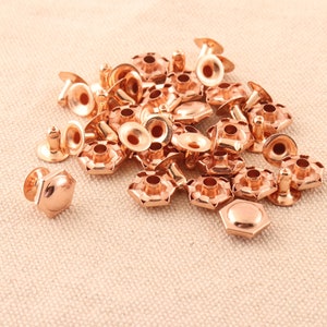 May include: A pile of rose gold metal rivets with a hexagonal shape and a raised center. The rivets are scattered on a beige fabric surface.