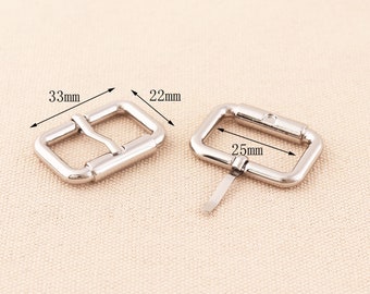 Könnte beinhalten: Zwei silberne Gürtelschnallen aus Metall. Die größere Schnalle ist 25 mm breit, die kleinere Schnalle 33 mm lang und 22 mm breit.