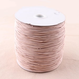 Può includere: Un rotolo di cordoncino elastico rosa chiaro avvolto attorno a un rotolo di cartone bianco. Il cordoncino è liscio e sembra essere fatto di un materiale elastico.
