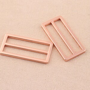 May include: Two rose gold metal slider buckles. Each buckle has two horizontal bars.