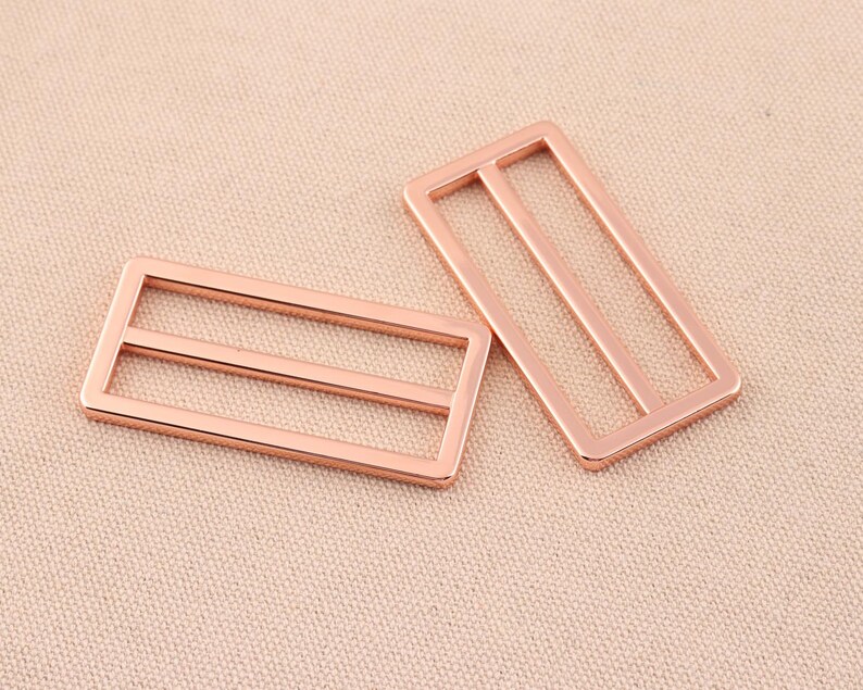 May include: Two rose gold metal slider buckles. Each buckle has two horizontal bars.