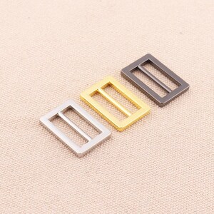 May include: Three metal slider buckles in different finishes: silver, gold, and black. The buckles are rectangular with two parallel slots.