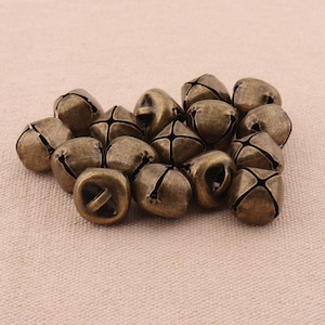 May include: A collection of small, antique brass-colored bells. Each bell has a cross-shaped slit design and a loop for hanging. The bells are clustered together on a neutral-toned fabric background, creating a rustic aesthetic.
