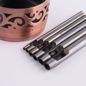 May include: A set of metal hole punches of varying sizes, arranged next to a copper-colored container with a decorative cutout design. The punches are cylindrical with a hollow center.