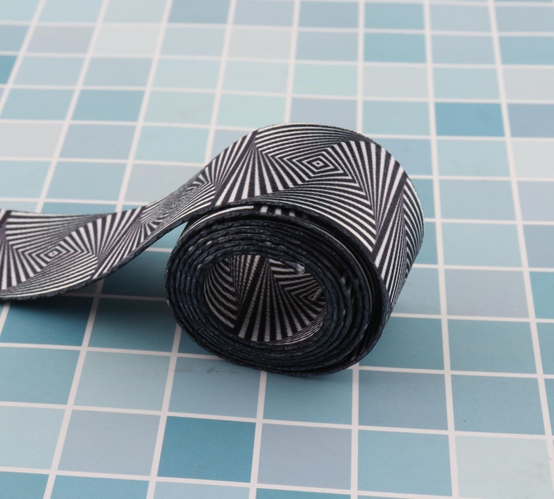 May include: A black and white patterned elastic band rolled up on a blue tiled surface. The pattern is a series of concentric circles with a diamond shape in the center.
