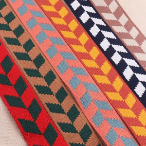 Op de afbeelding: Close-up van verschillende kleurrijke geweven banden met een herhalend chevronpatroon. De banden tonen een verscheidenheid aan kleuren, waaronder rood, groen, bruin, koraal, blauw, geel, marine en beige. De banden zijn waarschijnlijk voor tassen of andere accessoires.