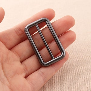 May include: A black metal rectangular buckle with two parallel bars inside. The buckle is designed for attaching straps or belts.