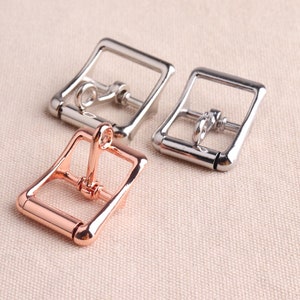 May include: Three metal belt buckles, one in silver, one in rose gold, and one in silver. The buckles are all the same size and shape, with a rectangular frame and a small loop on the top.