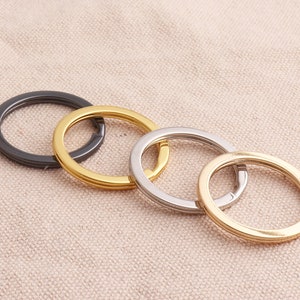 May include: Four key rings in different sizes and finishes: black, gold, silver, and gold. The key rings are arranged in a circle on a beige fabric background.