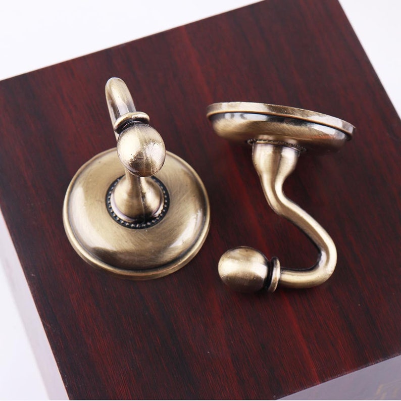 May include: Two antique gold-toned hooks with a decorative design. The hooks are mounted on a dark wood surface.