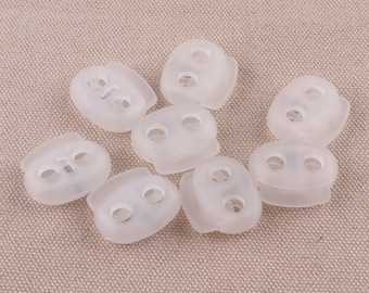 May include: Eight white plastic toggle clasps with two holes each. The clasps are arranged in a loose circle on a beige fabric surface.