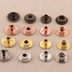 Puede incluir: Surtido de botones a presión metálicos en varios acabados, incluyendo oro, plata, bronce y cobre. Los botones a presión están dispuestos en filas sobre un fondo de tela marrón claro.