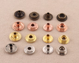 May include: Assortment of metal snap fasteners in various finishes, including gold, silver, bronze, and copper. The snaps are arranged in rows on a light brown fabric background.