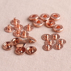 Könnte beinhalten: Eine Sammlung von roségoldenen Metall-Druckknöpfen, einige mit erhabener Mitte und einige mit flachem Oberteil. Die Knöpfe sind in einem zufälligen Muster auf einer beigefarbenen Stoffoberfläche angeordnet.