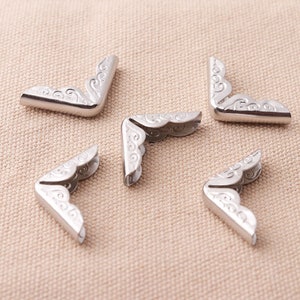 May include: Five silver metal corner protectors with a decorative, scalloped edge. The protectors are designed to protect the corners of books, journals, or other items.