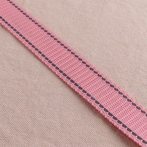 May include: A close-up of a pink fabric strap with black stitching. The strap is on a beige fabric background.