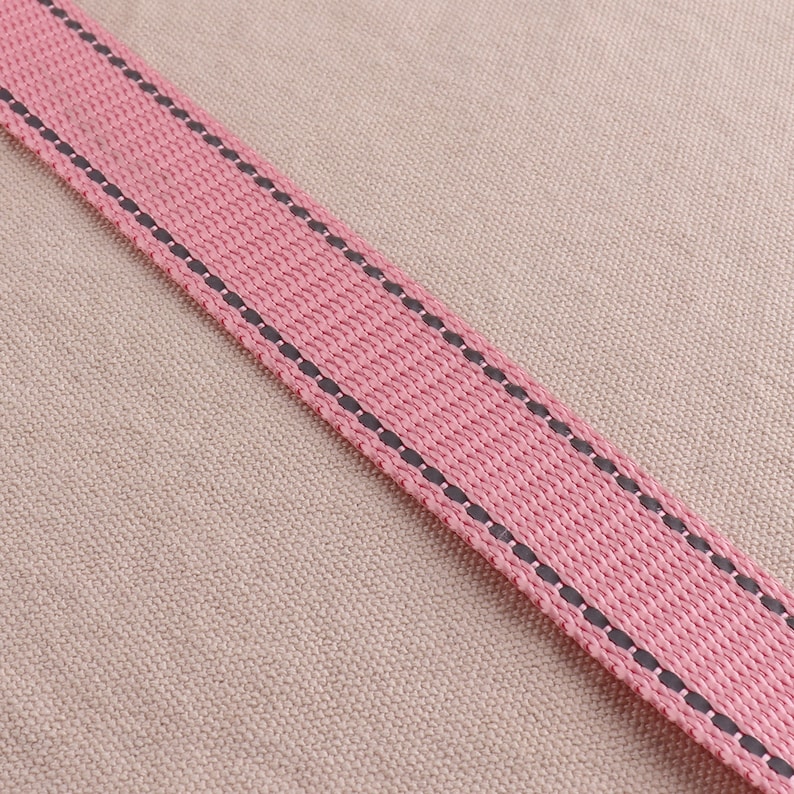 May include: A close-up of a pink fabric strap with black stitching. The strap is on a beige fabric background.