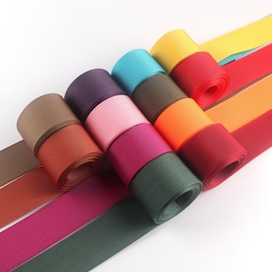 Puede incluir: Una colección de rollos de cincha de nailon de colores, en tonos marrón, naranja, rosa, morado, turquesa, verde, amarillo, rojo y naranja. Los rollos están apilados, con algunos desenrollados para mostrar la cincha plana.