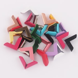 May include: Assortment of colorful metal corner protectors in various colors, including pink, blue, green, black, and gold. These corner protectors are ideal for scrapbooking, photo albums, and other craft projects.
