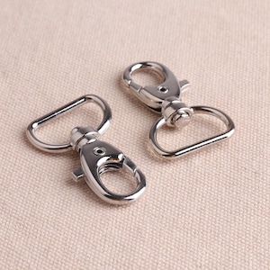 May include: Two silver metal swivel clasps with a D-ring on one side and a hook on the other. The clasps are commonly used for attaching straps, leashes, or other accessories.