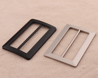 May include: Two metal belt buckles, one black and one silver. Both buckles are rectangular with a single slot in the center.