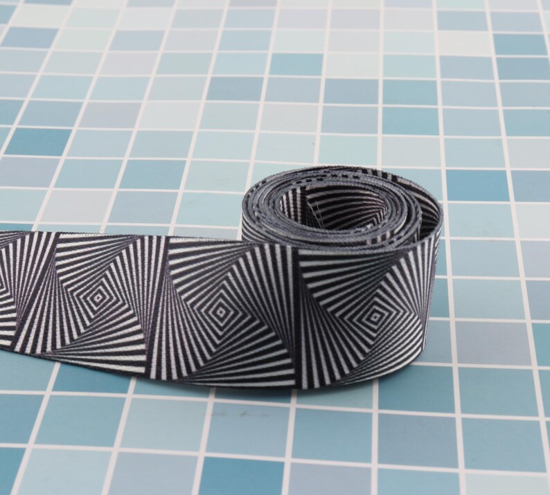 May include: A black and white patterned ribbon with a swirling, optical illusion design. The ribbon is rolled up and is on a blue tiled surface.