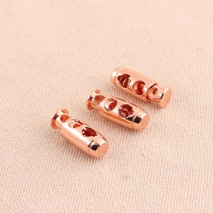 Puede incluir: Tres topes de cordón de palanca de oro rosa. Cada tope tiene una forma cilíndrica con un agujero en el centro y dos agujeros más pequeños a cada lado.