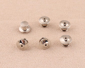 Op de afbeelding: Zes zilveren metalen pinruggen om badges of andere items aan kleding te bevestigen. De pinruggen hebben een ronde, platte basis met een klein gaatje in het midden.